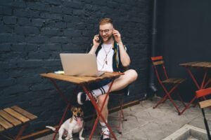 Cheerful digital nomad with laptop talking on mobile phone in sidewalk cafe