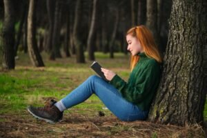 Redhead young woman reading a book sitting under tree in forest.