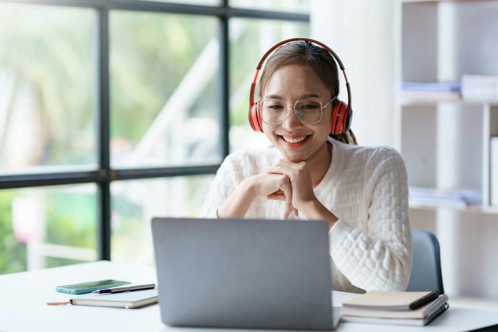Cute young Asian woman wearing headphones listening to music happily on her laptop enjoying listen