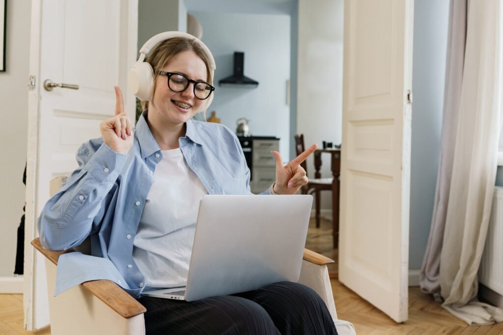 A cheerful woman enjoys her music, giving a peace sign while working on her laptop in a cozy room.